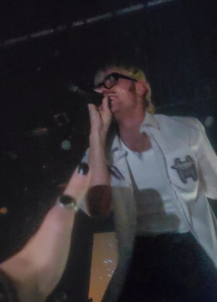 a man in white shirt and black pants on stage with his hand up to the microphone