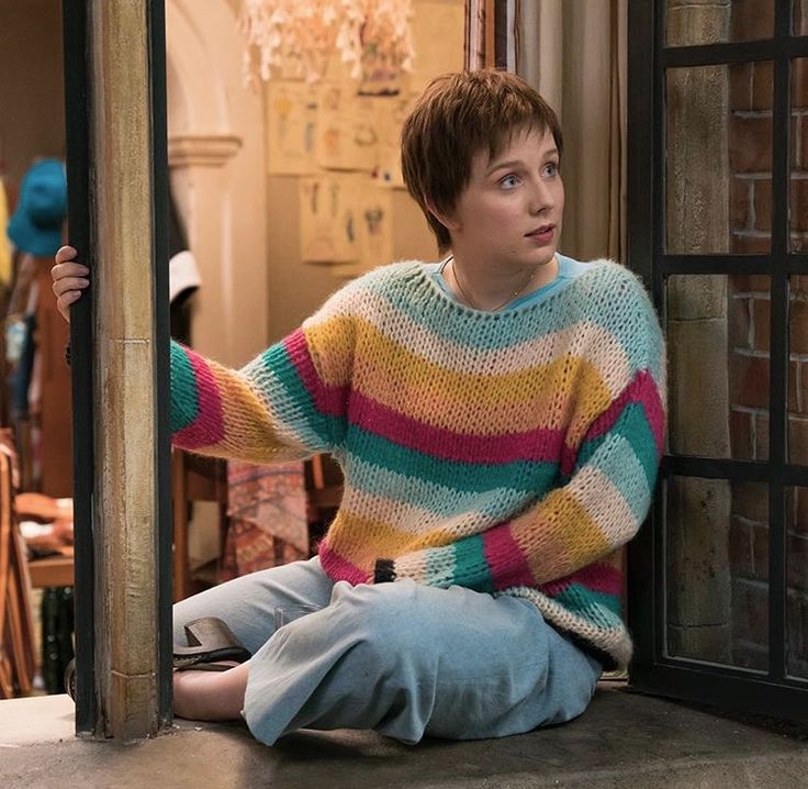 a young boy sitting on the window sill in front of a brick wall wearing a multicolored sweater