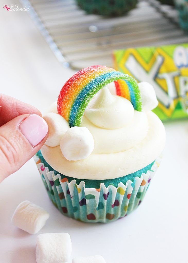 a hand holding a rainbow cupcake with marshmallows on the table next to it