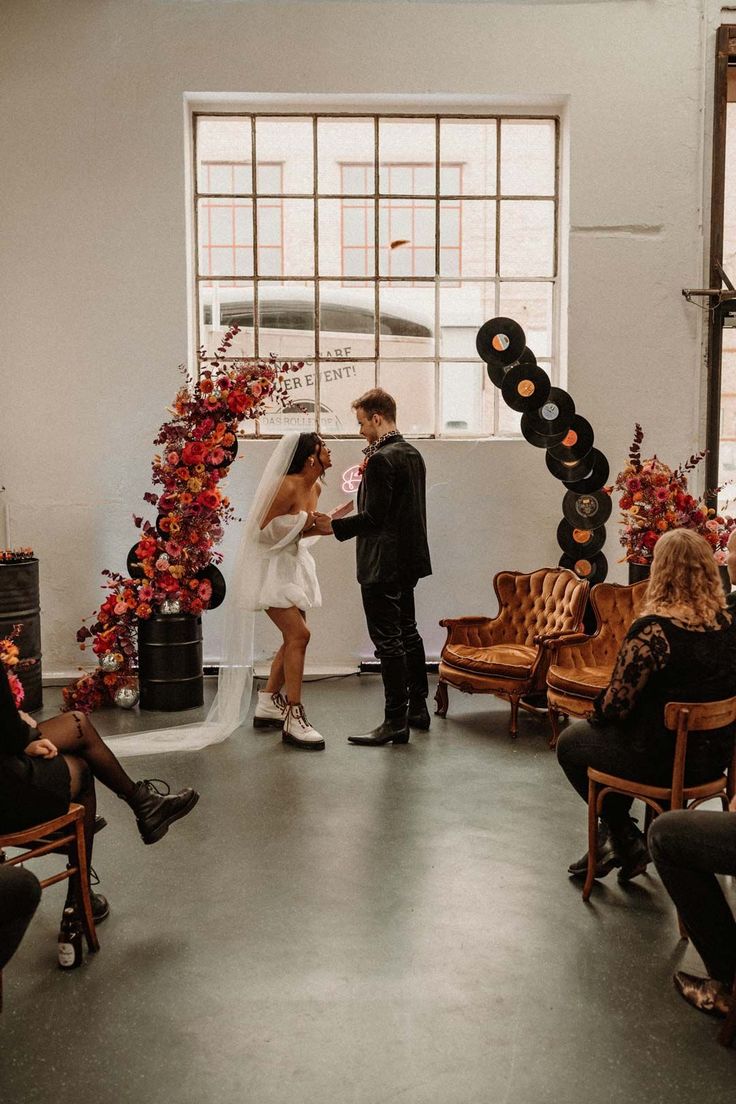 a bride and groom are standing in front of an arch with flowers on it, surrounded by chairs