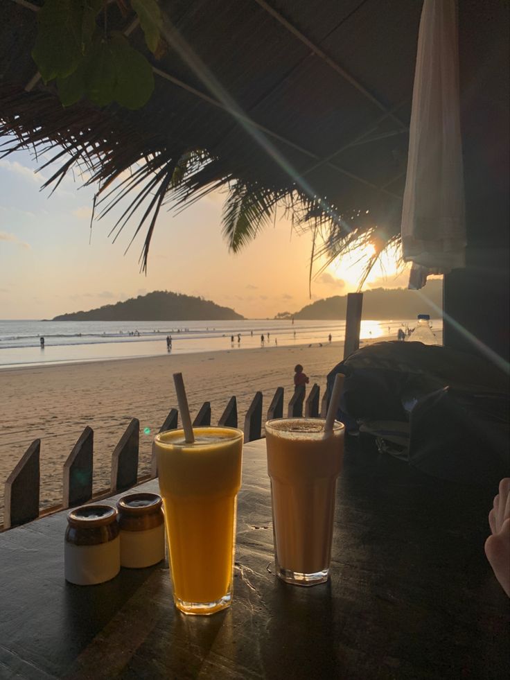 two drinks sitting on top of a wooden table next to the ocean with people in the background