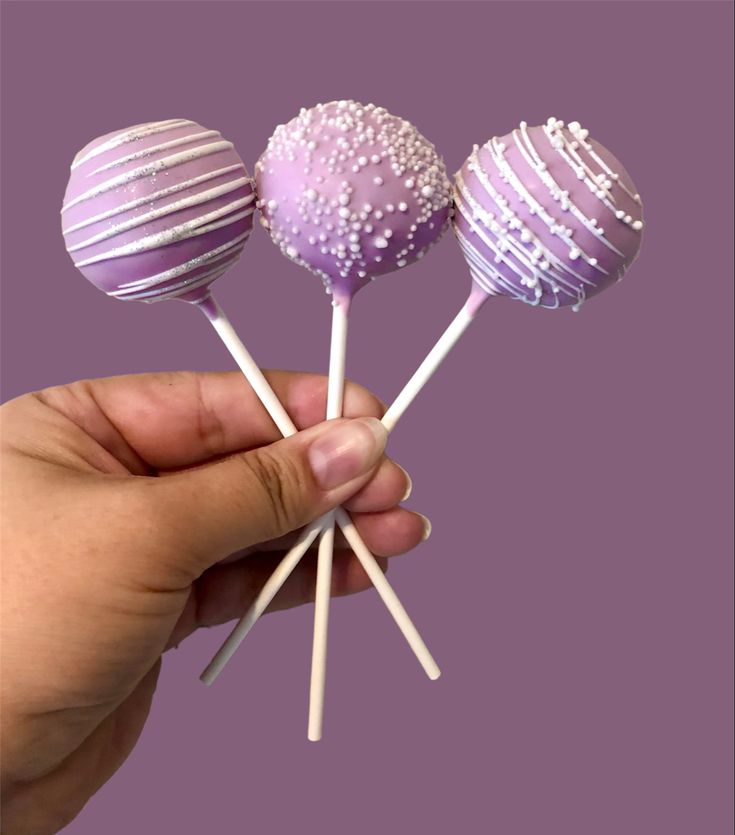 a hand holding three purple cake pops with white sprinkles on them in front of a purple background