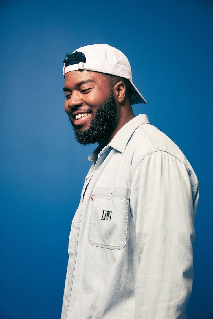 a man with a beard wearing a white hat and smiling at the camera while standing against a blue background