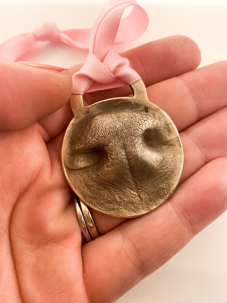 a hand holding a gold medal with a pink ribbon on it's end and a ring in the middle