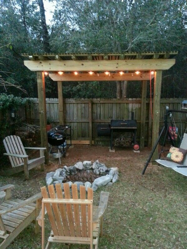 a fire pit surrounded by chairs and lights in a backyard with a grill on the other side