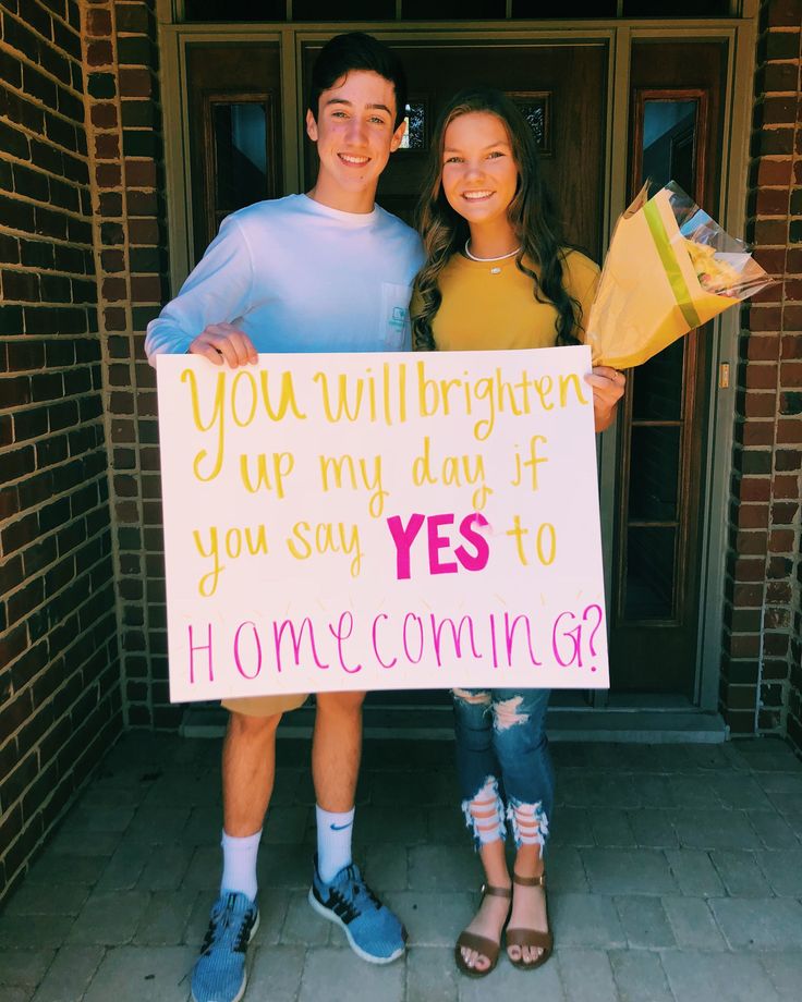 two people standing in front of a door holding a sign that says you will brighten up my day if you say yes to home coming?