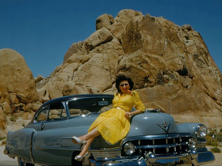 a woman in yellow dress sitting on the hood of an old car with rocks behind her