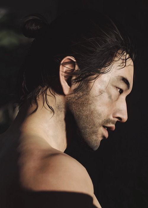 a man with long hair and no shirt looking off to the side, in front of a black background