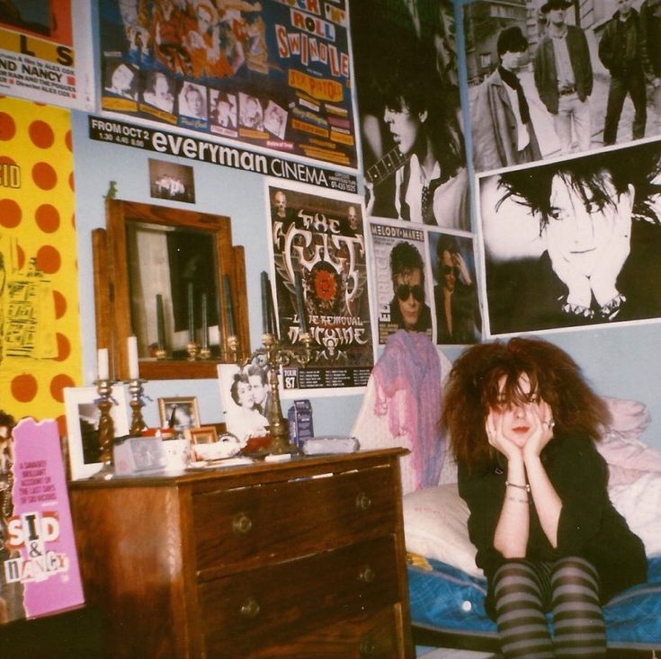 a woman sitting on top of a bed next to a dresser and wall covered in posters