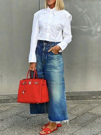 a woman standing in front of a wall holding a red handbag and wearing blue jeans