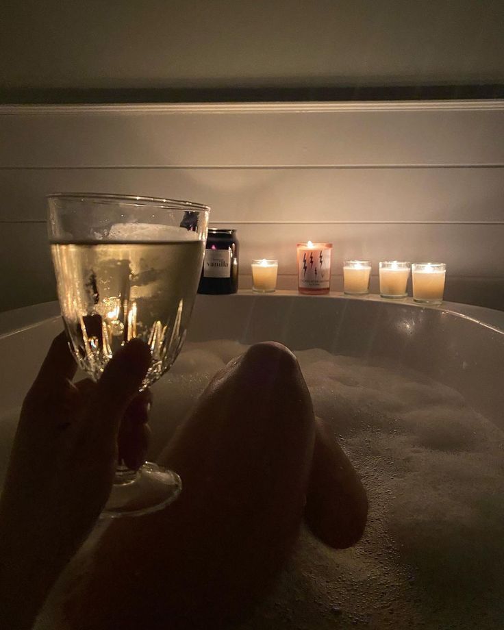 a person sitting in a bath tub holding a wine glass with candles around it and on the side