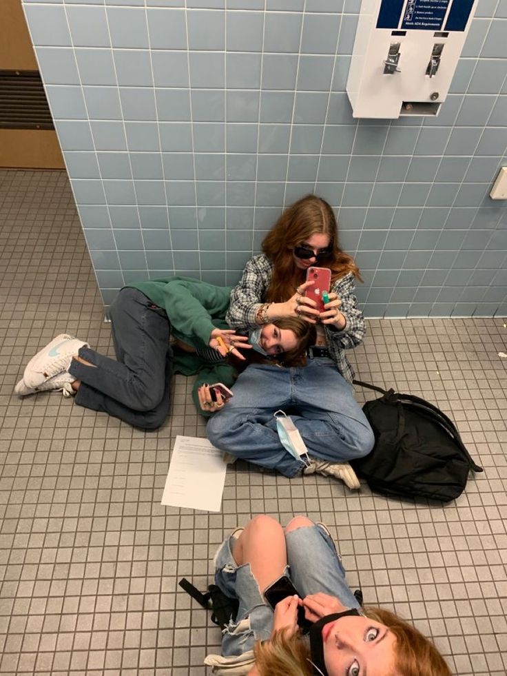 two women sitting on the floor using their cell phones in a public bathroom with tiled walls