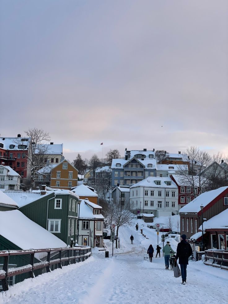 people walking down a snow covered road in the middle of some small town with lots of buildings