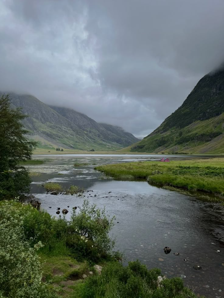 a river running through a lush green valley under a cloudy sky with mountains in the background