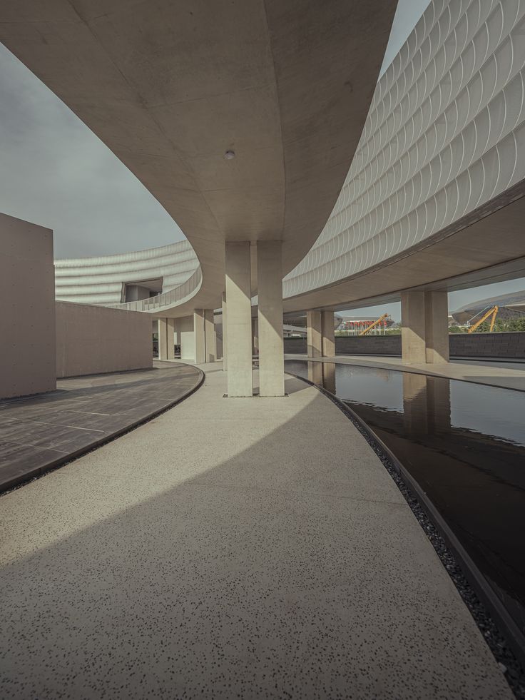 an empty walkway next to a body of water in front of a building with curved roof