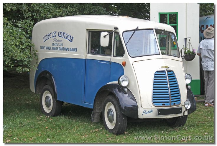 an old blue and white truck is parked in the grass with people looking at it