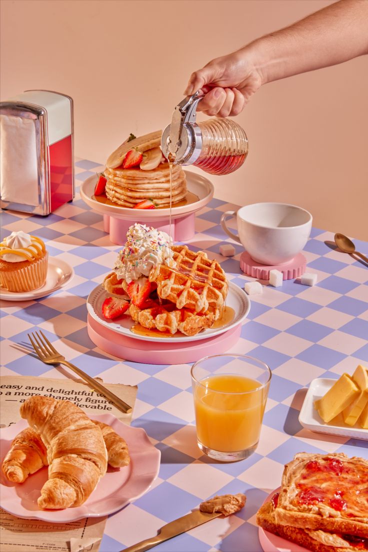 a person pouring syrup on some waffles at a table with other breakfast foods