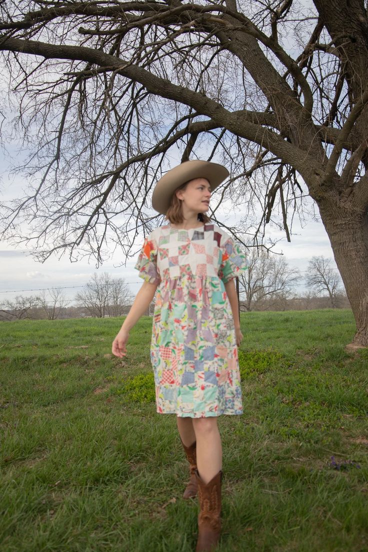 a woman in a dress and cowboy hat walks through the grass under a large tree