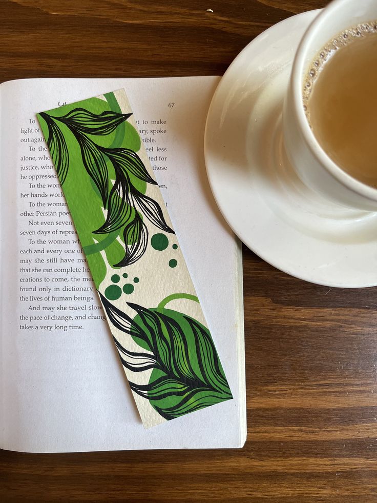 a bookmark with green leaves on it next to a cup of tea and saucer