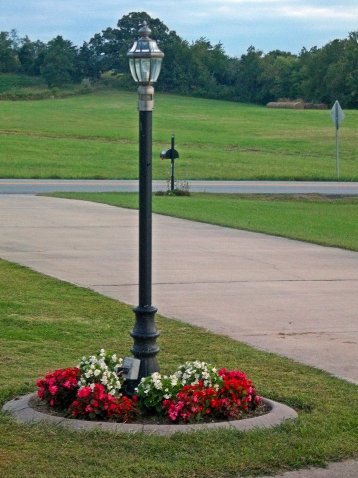 a lamp post with flowers around it in the middle of a sidewalk and grass area