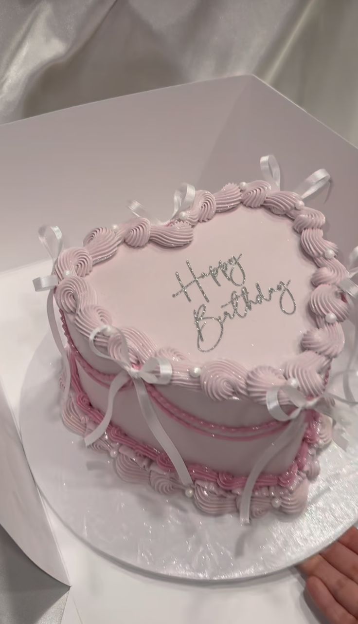 a pink and white birthday cake sitting on top of a table next to a person's hand