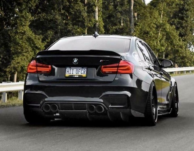 the rear end of a black bmw car on a road with trees in the background