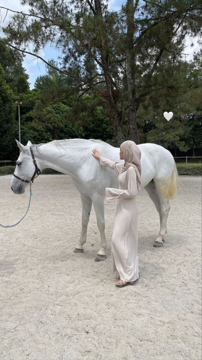 a woman standing next to a white horse on top of a sandy field with trees in the background