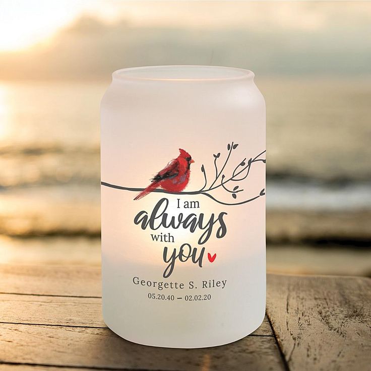 a candle with a cardinal on it sitting on a wooden table near the water at sunset