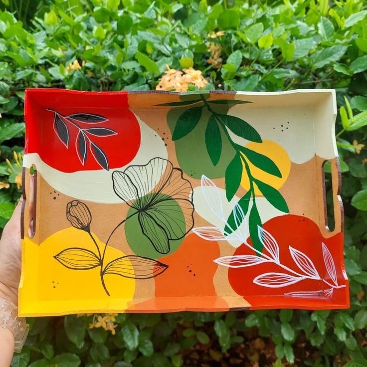 a person holding up a painted tray in front of some bushes and trees with green leaves on it