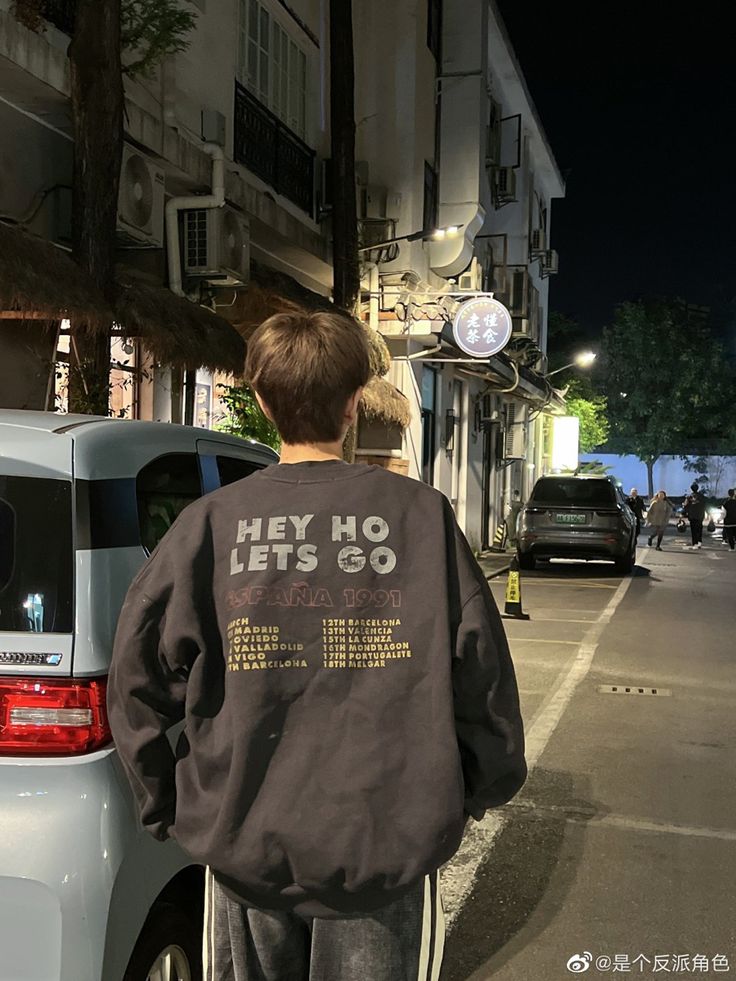 a boy walking down the street with his back turned to the camera, wearing a jacket that says hey no lets go