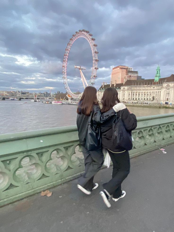 two women walking across a bridge next to the water with a ferris wheel in the background