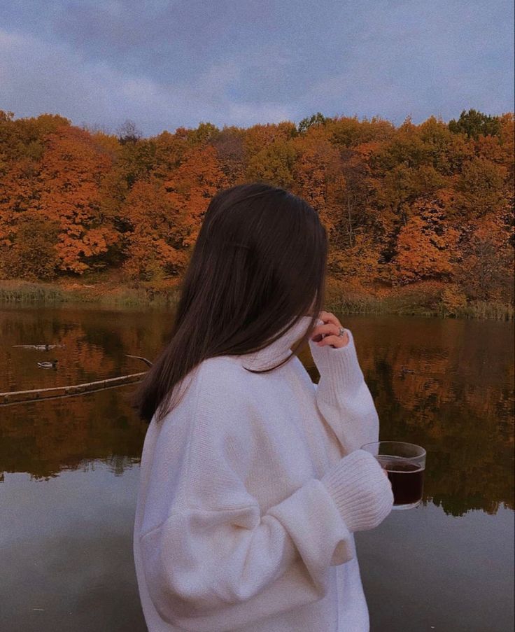 a woman standing in front of a body of water holding a cup and looking into the distance