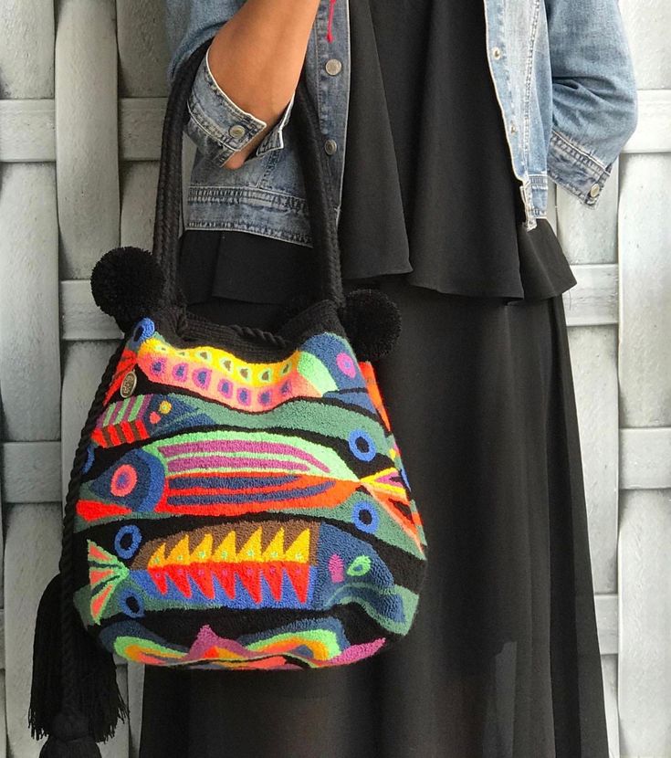 a woman is holding a colorful handbag in front of a white brick wall and wearing a denim jacket