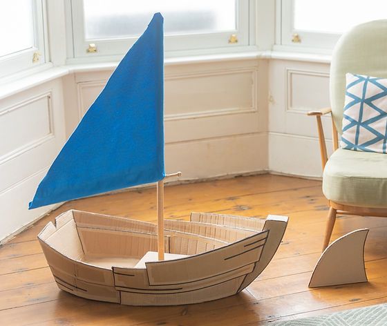 a cardboard boat sitting on top of a wooden floor next to a chair and window