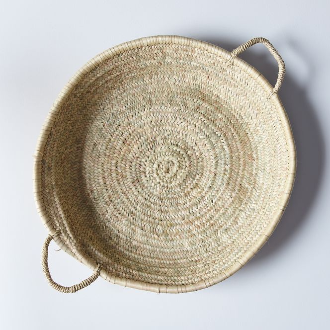 a round woven basket on a white surface with a string hanging from the bottom and two handles