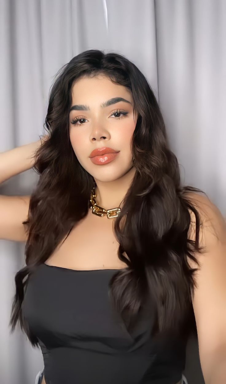 a woman with long dark hair wearing a black top and gold choker, standing in front of a curtain