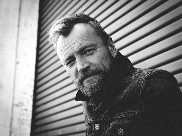 a black and white photo of a man with a beard looking at the camera while standing in front of a building