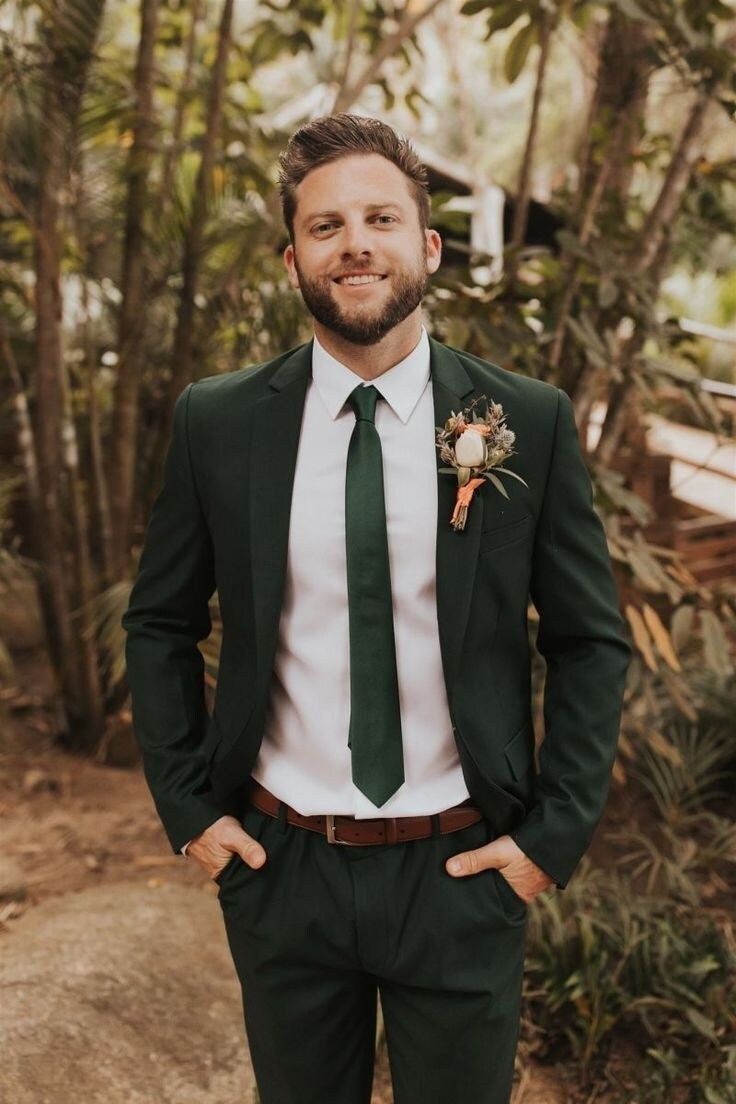 a man wearing a green suit and tie standing in front of some trees with his hands in his pockets