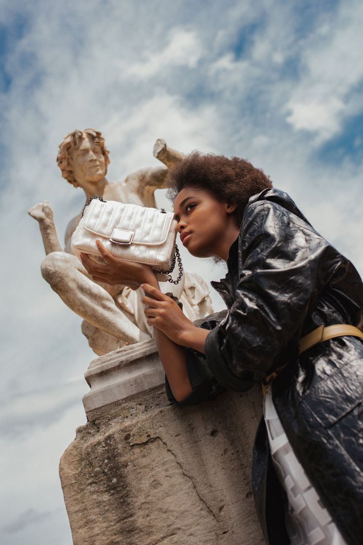 a woman leaning against a statue holding a white purse