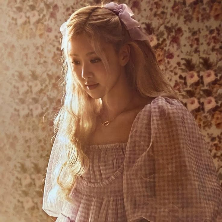 a woman with long blonde hair wearing a pink dress and sitting on a chair in front of a floral wallpaper