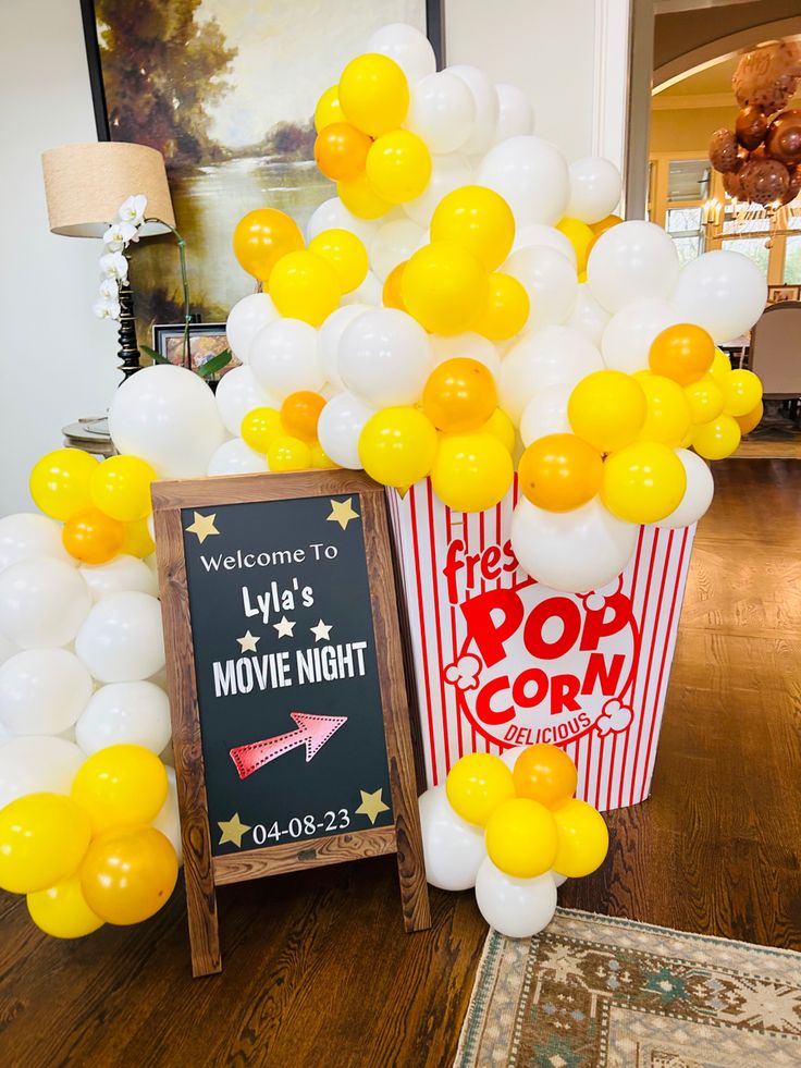 a popcorn bucket with yellow and white balloons next to a chalkboard sign that says movie night