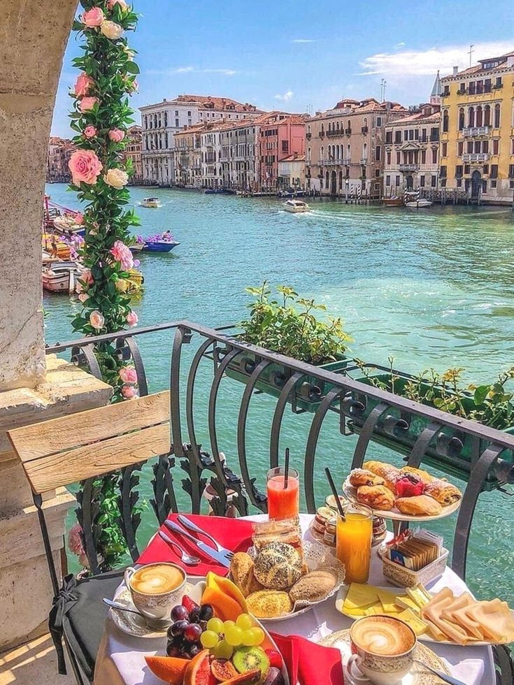 an outdoor table with food and drinks on it next to the water in venice, italy