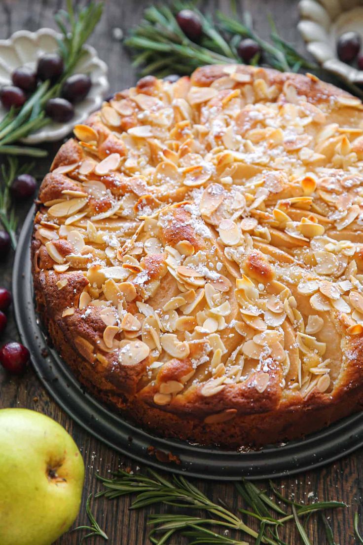 a cake with almonds on it sitting next to an apple and rosemary sprigs