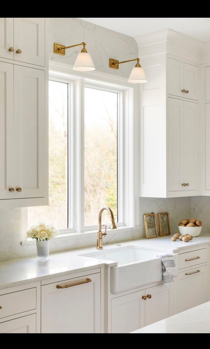 a kitchen with white cabinets and gold pulls on the faucet, sink, and window