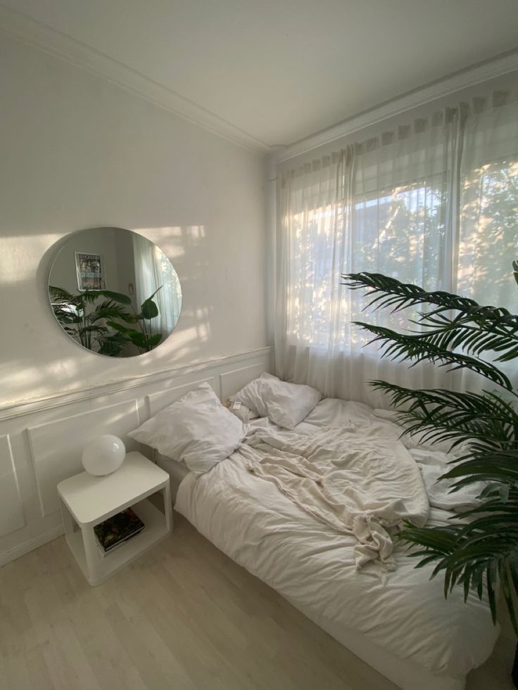 a white bed sitting under a mirror next to a plant in front of a window