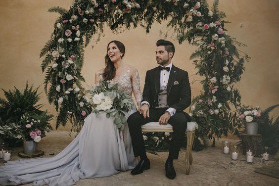 a man and woman sitting next to each other in front of an arch with flowers