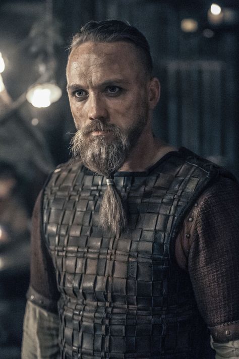 a man with a long beard wearing a leather vest and standing in a dark room