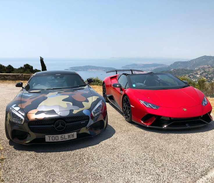 two sports cars parked next to each other in front of a mountain range with the ocean behind them