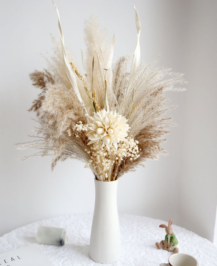 a white vase filled with dried flowers on top of a table next to a cup and saucer