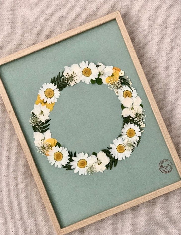 a wreath made out of daisies and greenery sits in a frame on the floor